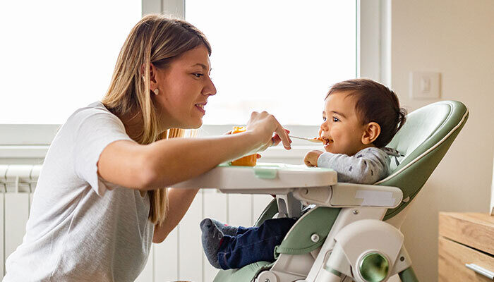 Young mother spoon feeding baby who is seated in a high chair in the living room