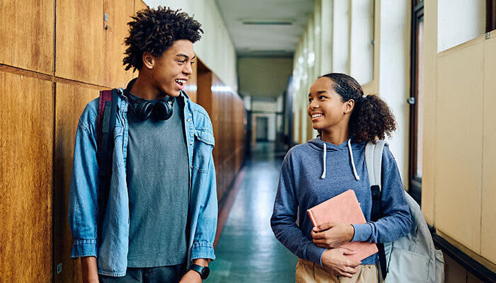 Happy Black teenage girl and her classmate communicating while walking through hallway at high school.