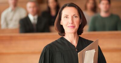 whhite woman in a black robe, with people out of focus seated behind her. She is holding papers.